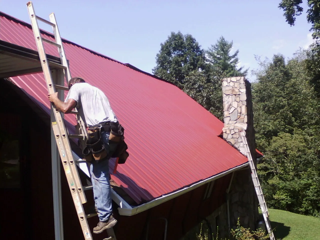 Roofer on a ladder beside a red metal roof and stone chimney — Soffit & Fascia Repair by Roofing Brothers in Henning